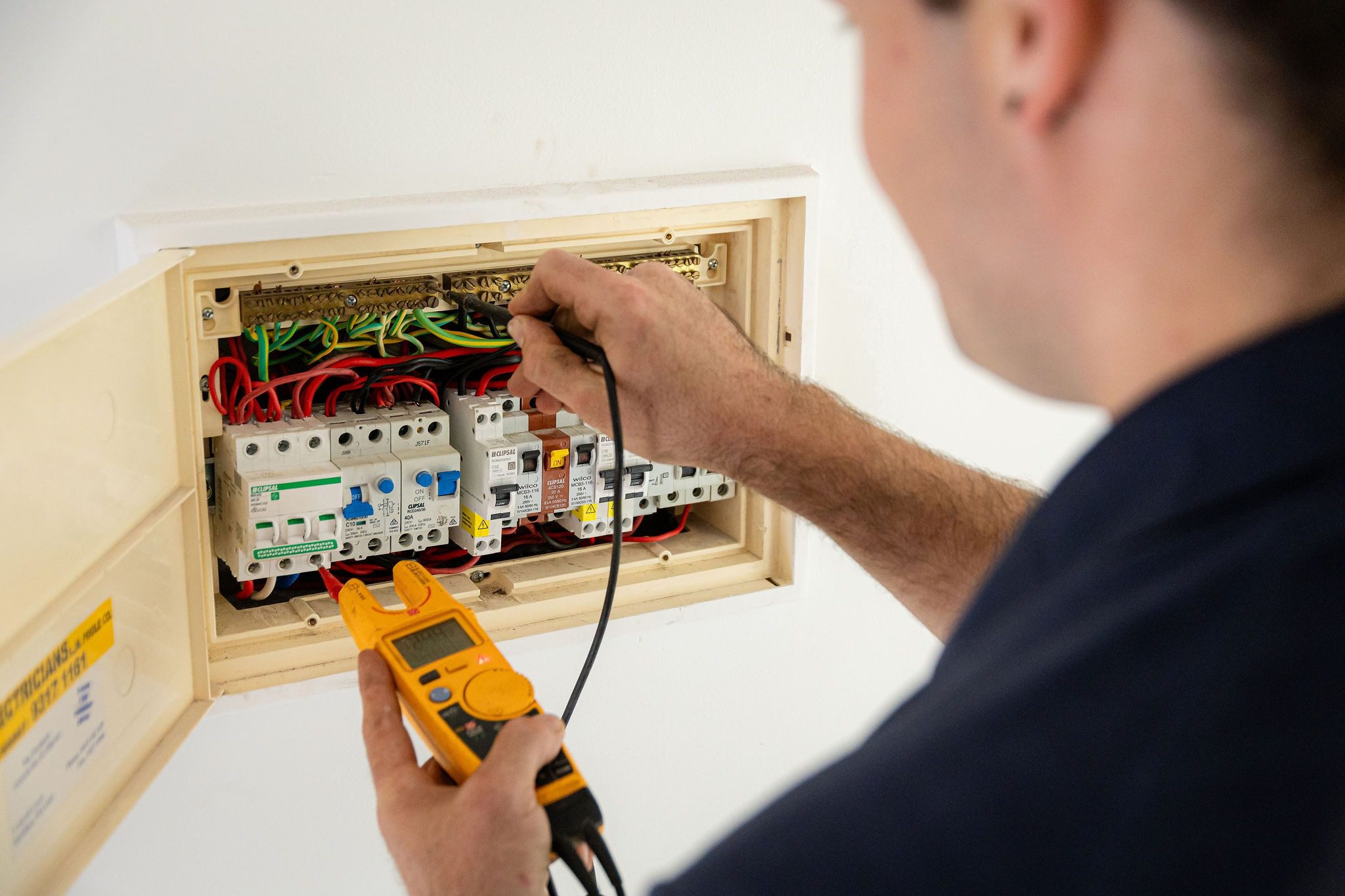 An electrician uses a yellow multimeter to test an open electrical panel with circuit breakers and wiring.