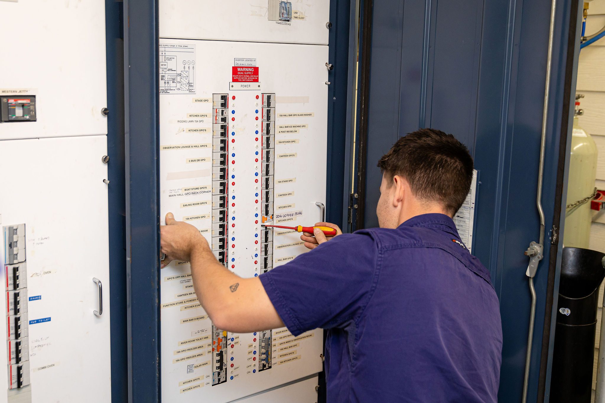 An electrician in Perth wearing a navy-blue uniform works on a large electrical panel, using a screwdriver to adjust circuit breakers.