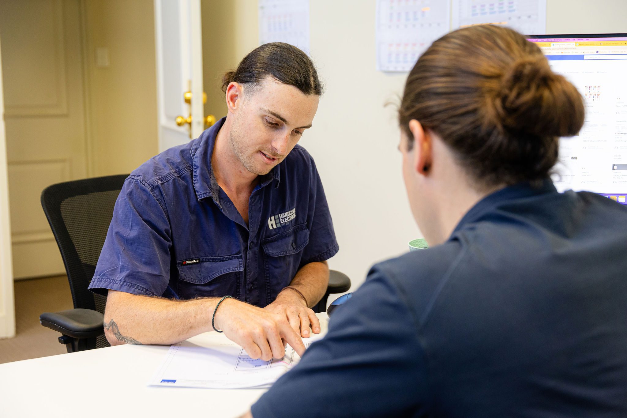 An electrician in Perth wearing a navy-blue uniform discusses electrical plans with a colleague in an office, pointing at documents on the table.